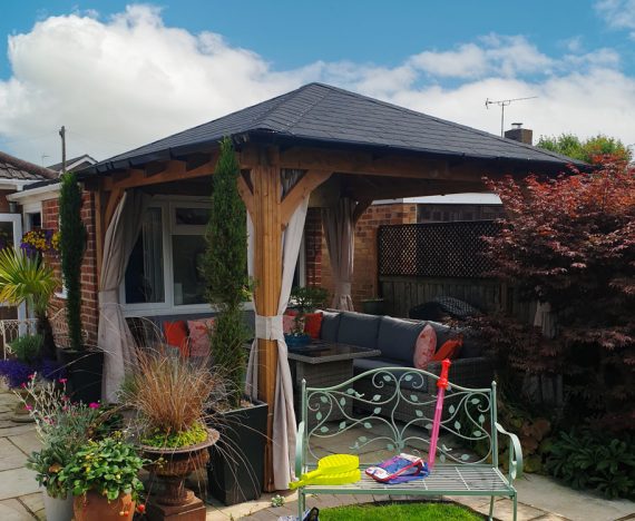 A wooden gazebo (Leviathan 3x3 curtain open gazebo) with a grey shingled roof stands in a garden setting. The gazebo features cream curtains and a seating area with cushions. Potted plants and a metal bench are nearby.