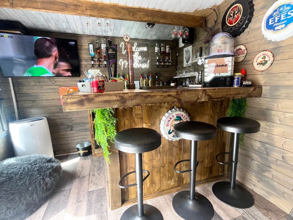 A small home bar in a log (Severn Log Cabin W5.0m x D3.0m) cabin with rustic wooden walls and décor. There is a wooden bar counter with stools, a TV, and various drinks and snacks.