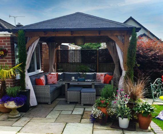 A wooden gazebo (Leviathan 3x3 curtain open gazebo) with a grey shingled roof stands in a garden setting. It features open sides with cream curtains, a comfortable outdoor seating area with cushions, and potted plants surrounding the space