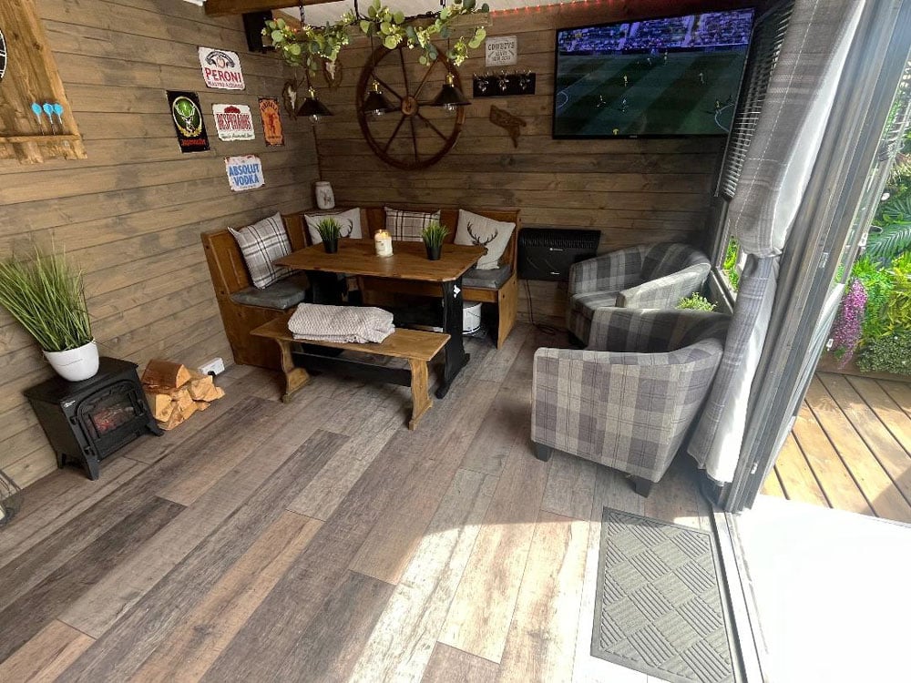  A cozy garden room (Severn Log Cabin W5.0m x D3.0m) with rustic wooden walls, a wood-burning stove, and a comfortable seating area with a table and chairs. A TV is showing a football match.